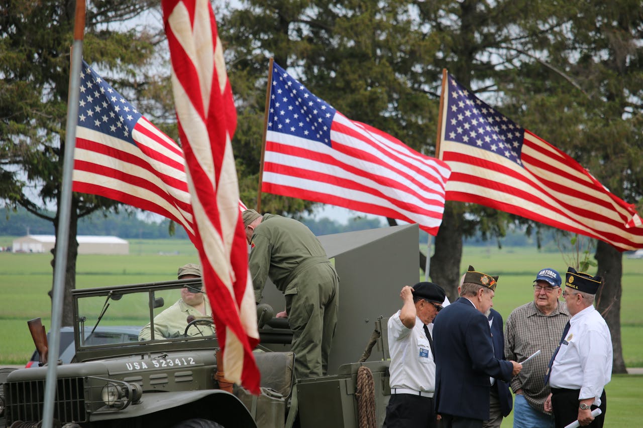 Group of veterans and soldiers by military vehicle with American flags in the background, symbolizing patriotism.